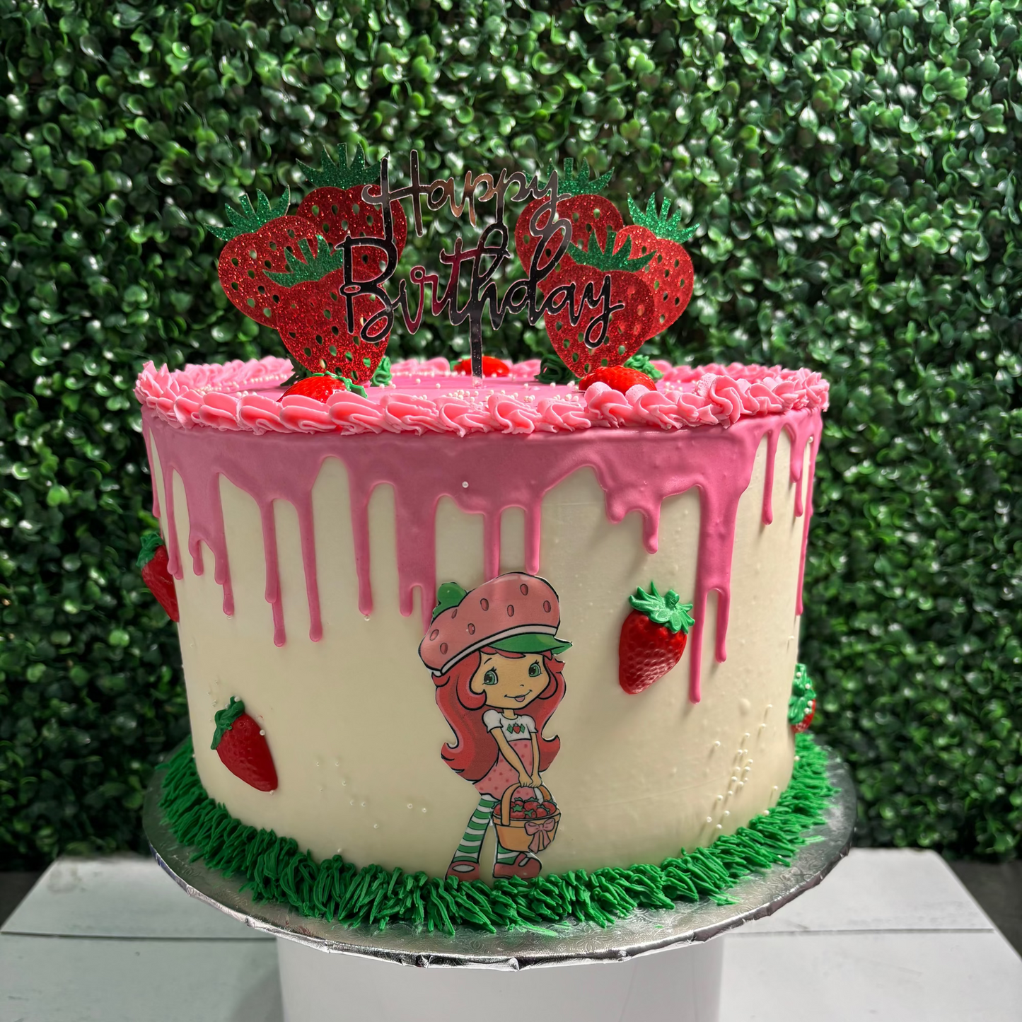 Birthday cake with pink frosting, strawberries, and Strawberry Shortcake character on a green leafy background