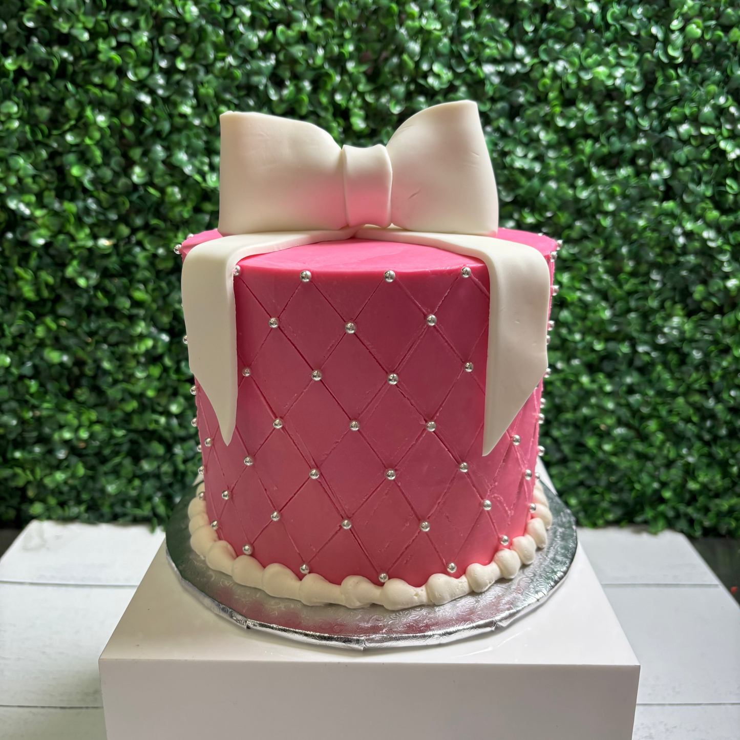 Pink cake with a white bow on a white stand against a green leafy background