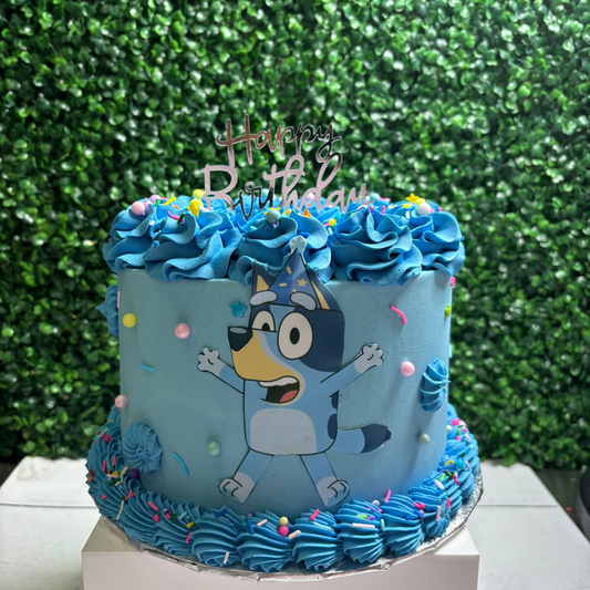 Blue birthday cake with Bluey cartoon character and 'Happy Birthday' topper against a green leafy background