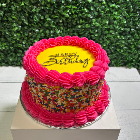 Birthday cake with pink frosting and colorful sprinkles on a white stand against a green leafy background